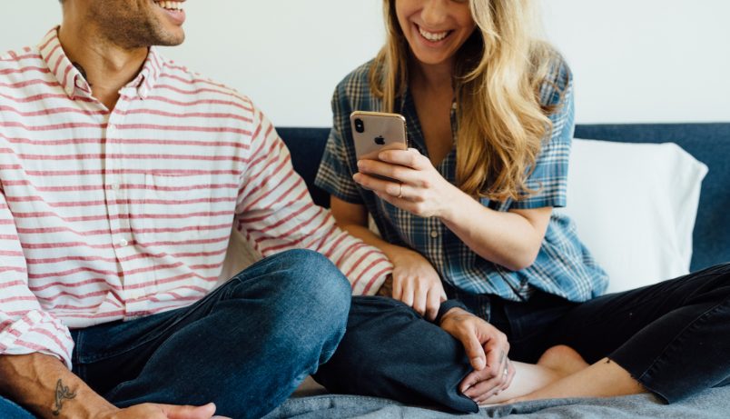 A man and woman sit cross-legged together on a bed, affectionately. The woman smiles at her phone in hand.